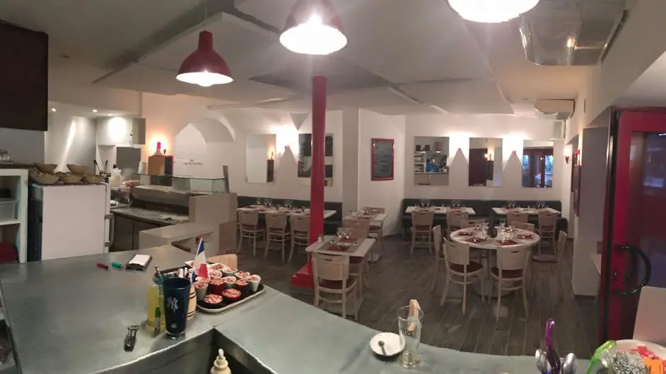 La salle : murs blancs, comptoir en zinc, poteau rouge et tables dressées